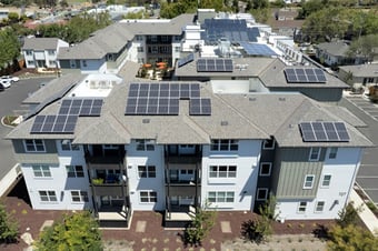 A four-story housing complex with white siding and dark gray balconies. Solar panels cover the light gray shingled roof. Bright umbrellas are visible in a center courtyard.