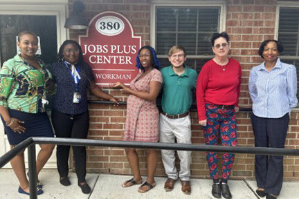 Six smartly dressed members of the RHHA Jobs Plus Program team in front of a Jobs Plus Center sign on a building