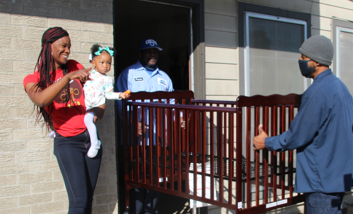 Mom and toddler smile at volunteers carrying a crib