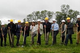 Eleven people in professional clothes and hard hats pose with shovels for a groundbreaking photo