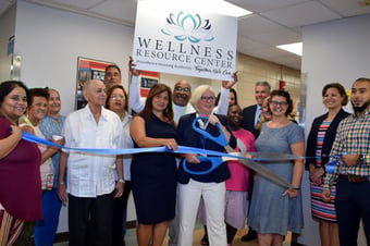 A crowd celebrates the opening of the Wellness Resource Center with a ribbon cutting. One participant holds up a sign with the WRC logo, and one holds a meter-long pair of novelty scissors up to the blue ribbon.