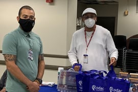 Two men in protective masks stand at a table bearing bags labeled "Codac"